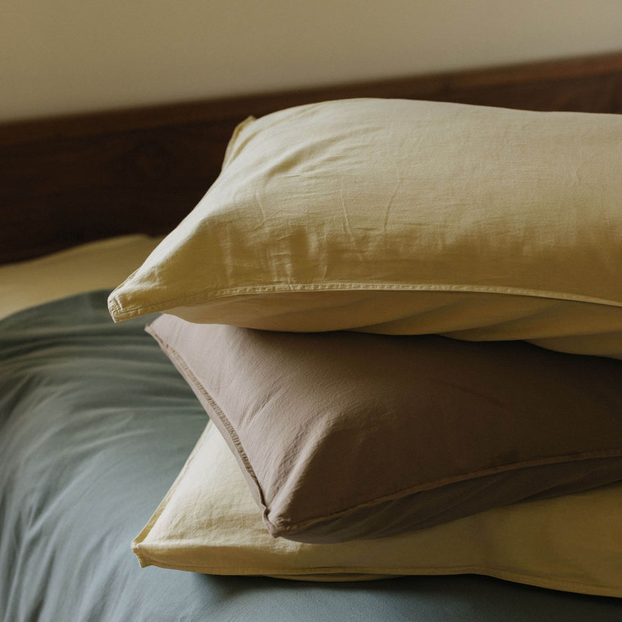 A stack of pillows in matte sateen colors Límon and Dune over a green bedcover on a wooden bed