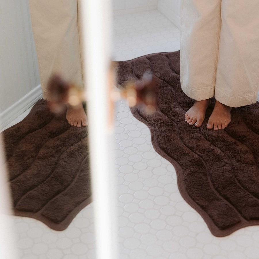 Brown bath mat on a white tiled bathroom floor with a person wearing beige pants.