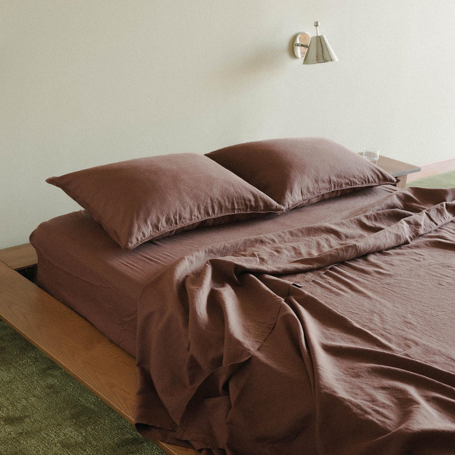 A close up on the wood bed with purple / brown sheets and two pillows. The flat sheet is pulled back a bit and there a chrome sconce above the bed