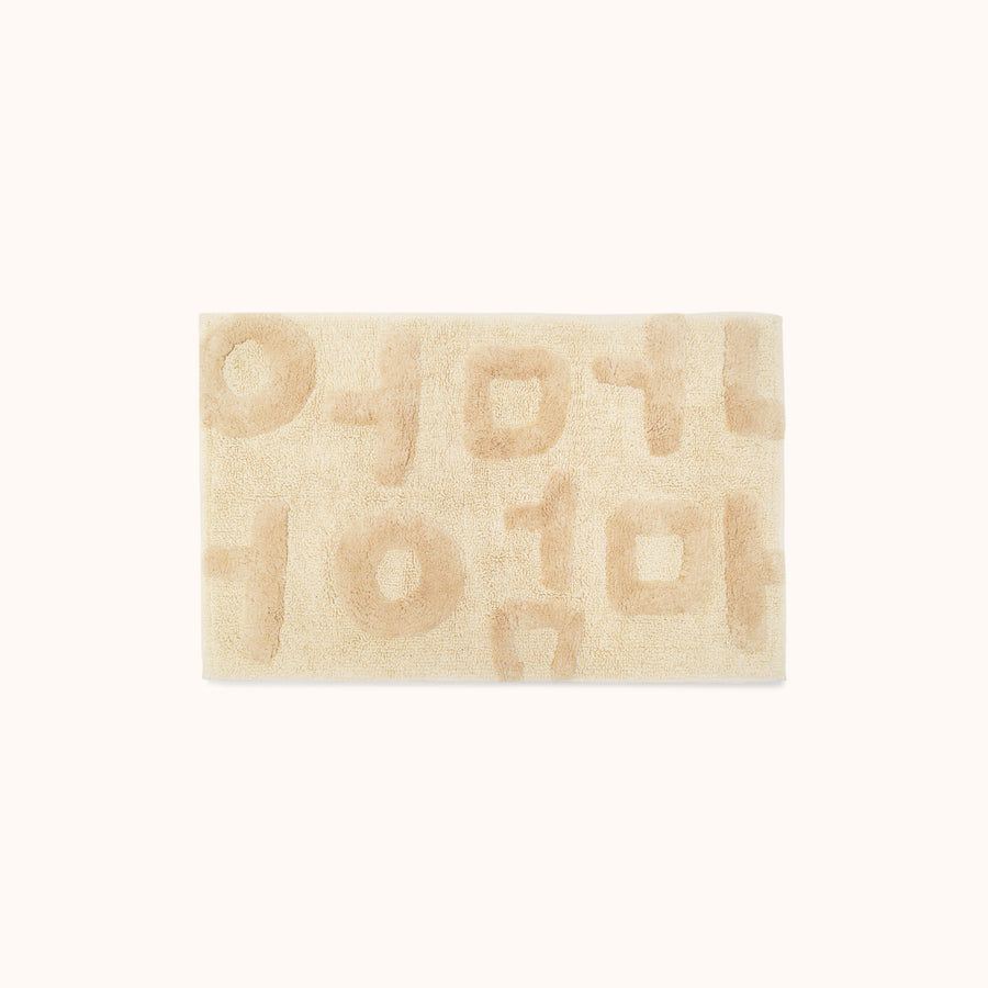 A rectangular beige bath mat with the word 'Mother' written in Korean characters, tufted in varying heights for a textured look.