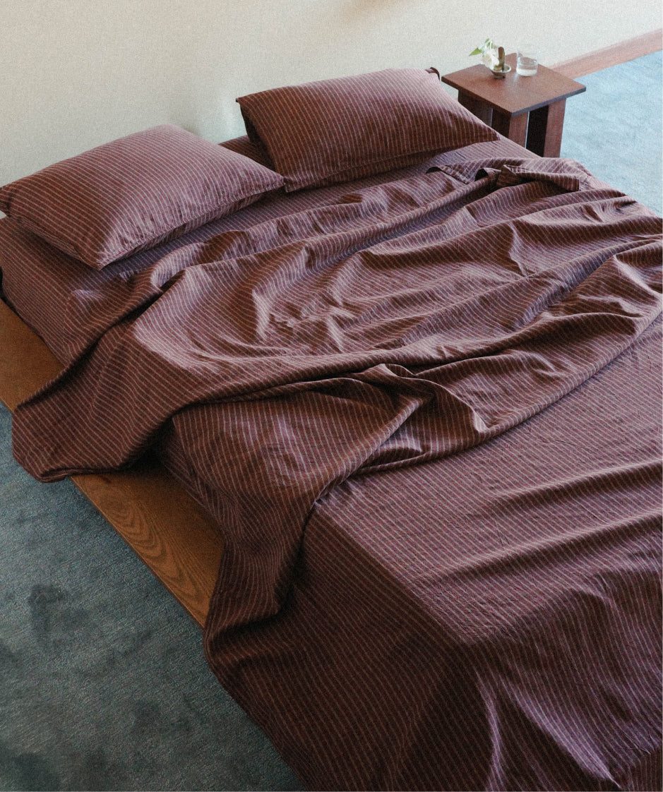 A bed dressed in maroon check bedding set with matching pillowcases, shown in a bedroom setting with a wooden nightstand and blue carpet.