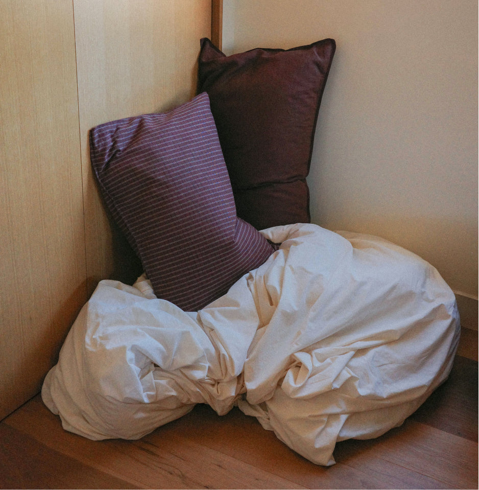 Two pillows, one dark plum and one maroon with a check pattern, resting on an off white comforter in a corner of a room with light wood walls and floor.