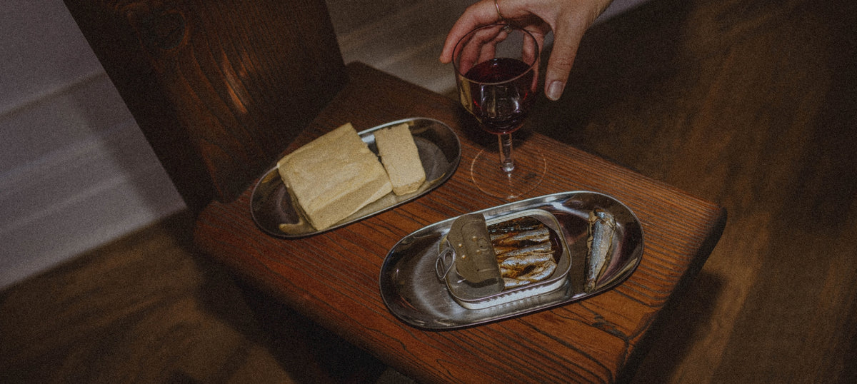 A hand holding a glass of red wine over a rustic wooden table, with two silver oval trays holding butter and tinned fish. The scene is dimly lit with a warm, cozy atmosphere.