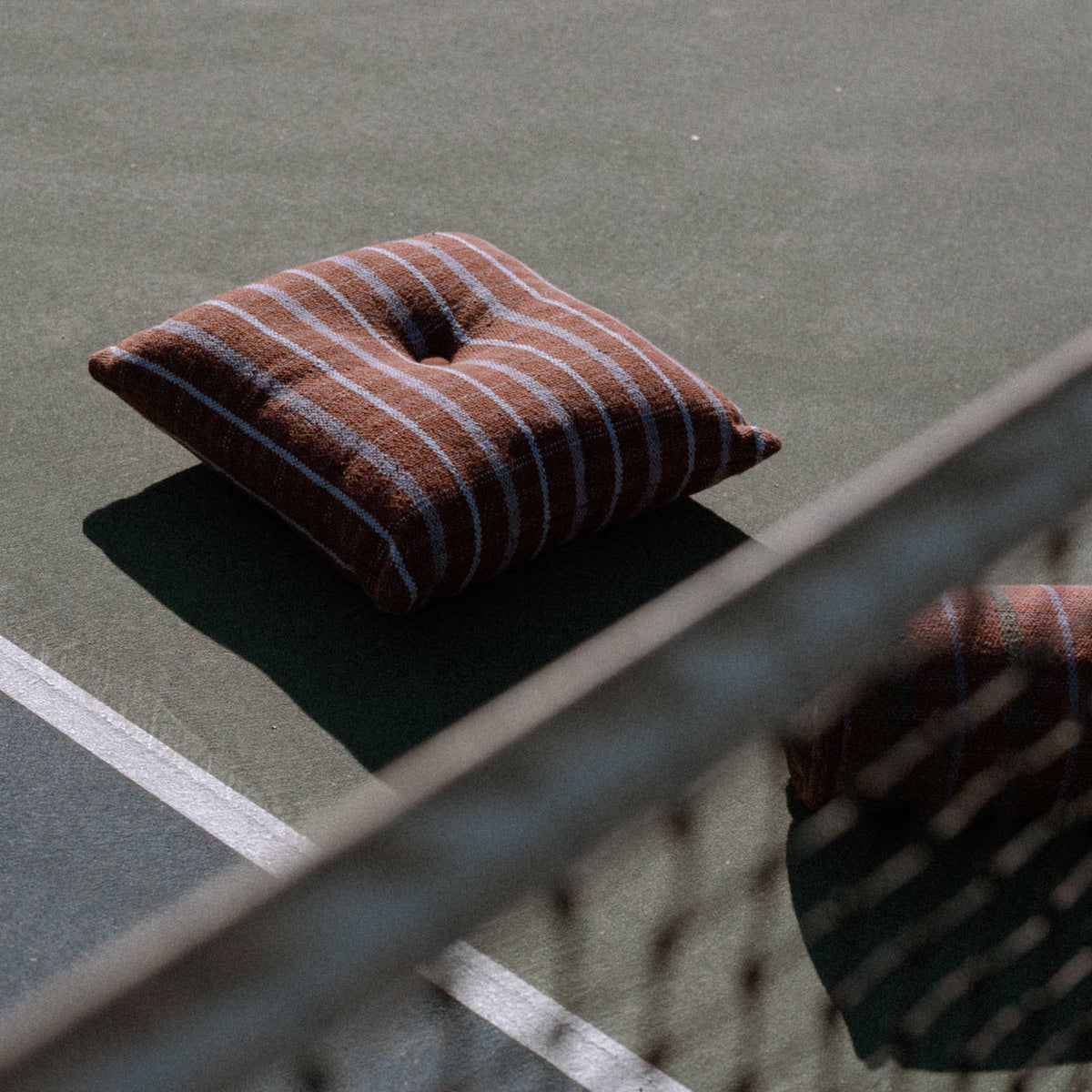 Brown and lavender check pillow with a button in the center on a tennis court with a blurred background.