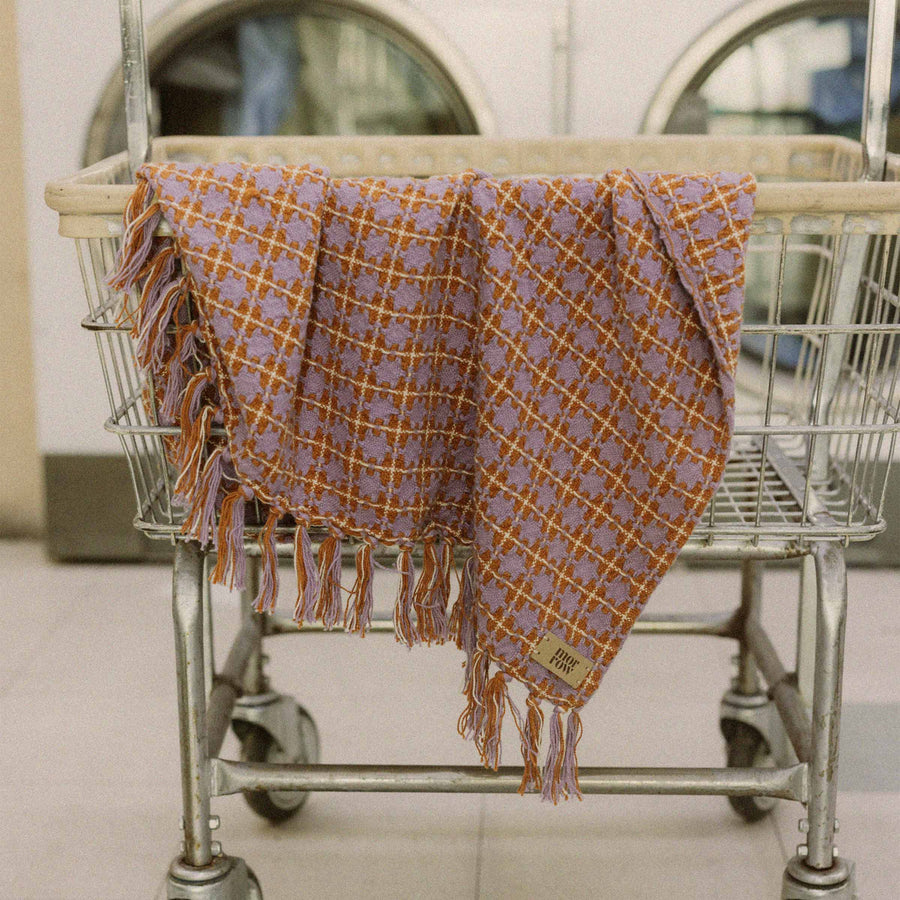 The Morrow Soft Goods Rosina blanket in a vintage Laundromat setting - the blanket is Rust and purple hued with tassels  