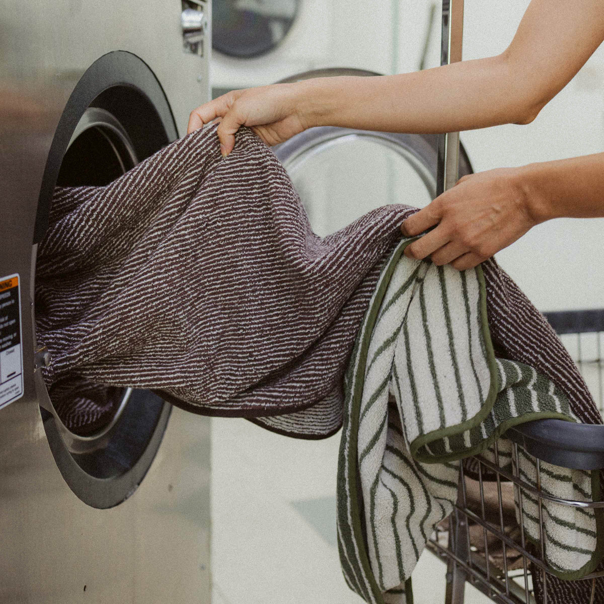 A brown and tan striped towel being pulled from a laundromat machine next to a laundry cart holding a green and tan striped towel. 