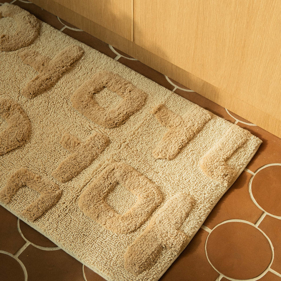 A rectangular beige bath mat with the word 'Mother' written in Korean characters, tufted in varying heights for a textured look on a terracotta tile floor.