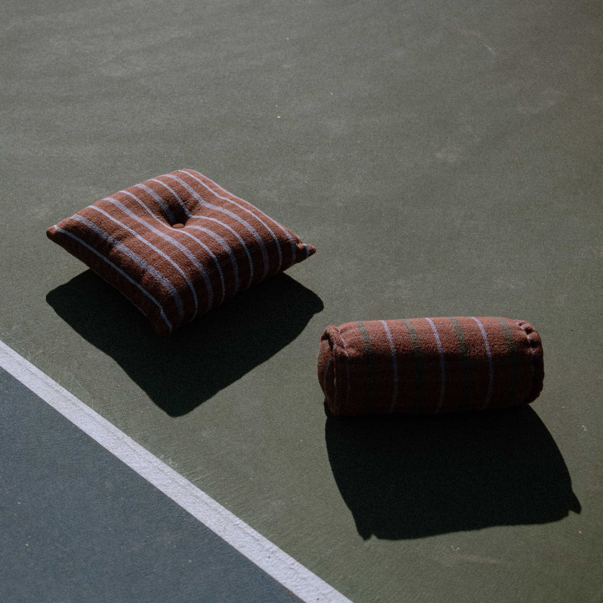 Brown and lavender striped pillow with a button in the middle and a Brown cylindrical bolster featuring green and lavender check pattern on a tennis court.