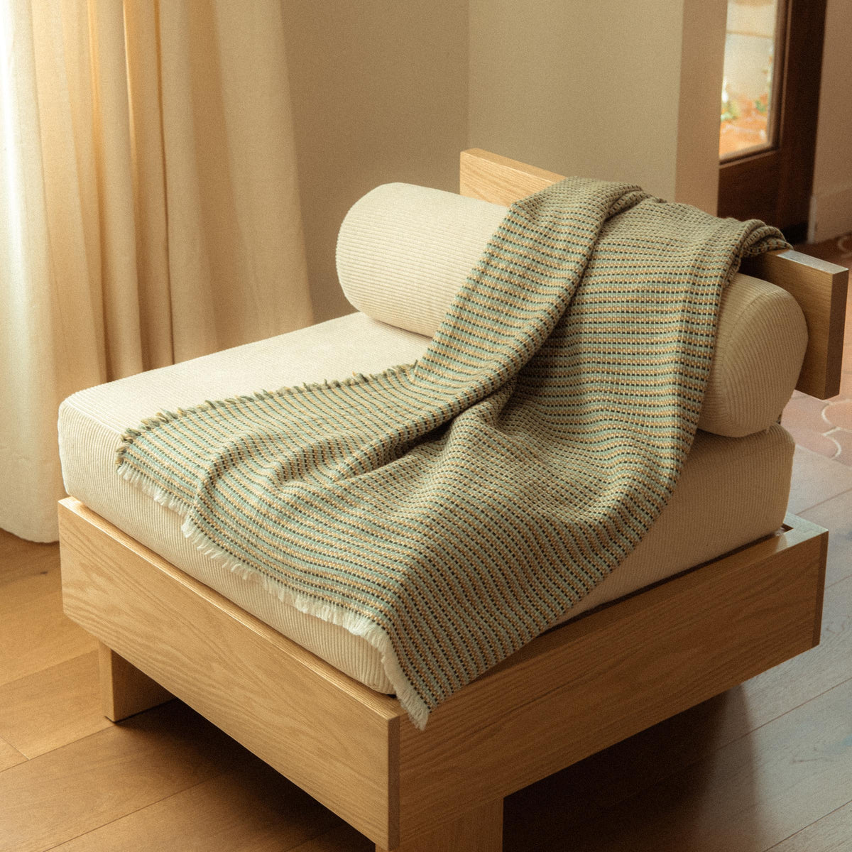 blanket with a subtle woven stripe pattern in golden yellow, blue, and green hues draped over a chair in a room with a curtain.