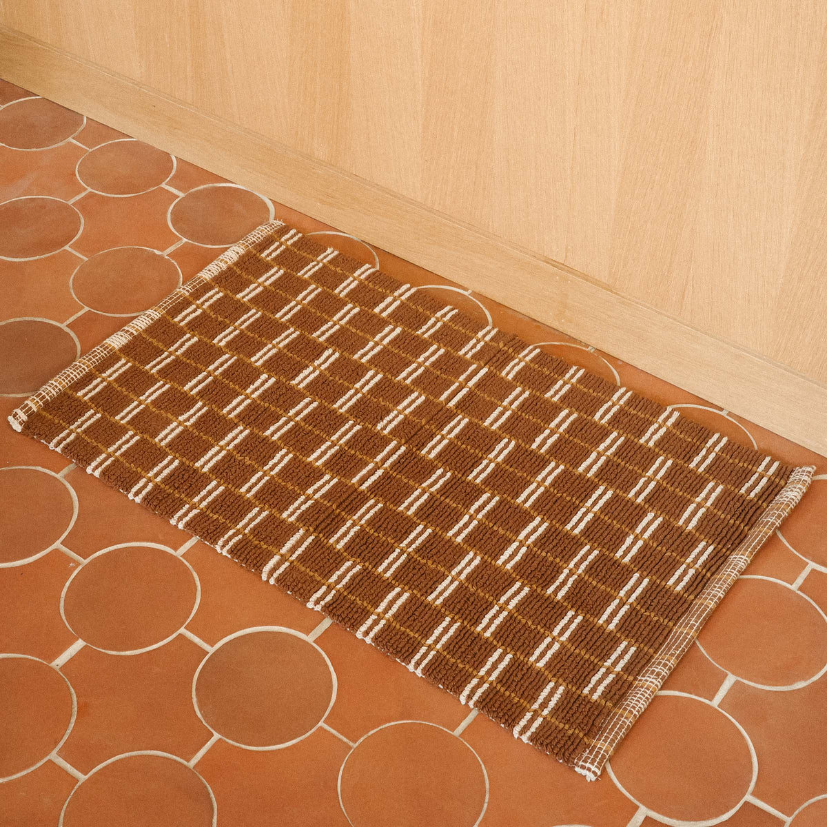 A brown mat with a tan geometric pattern on a terracotta tile floor. 
