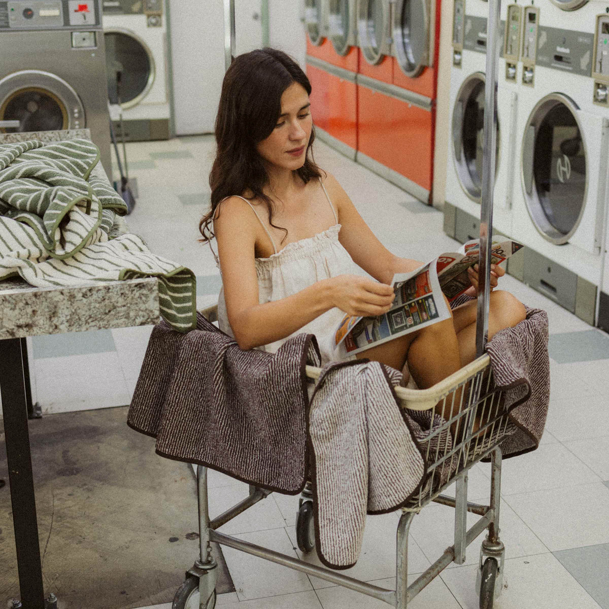 A woman sitting in a laundry cart in a laundromat reading a magazine surrounded by brown and tan striped towels with green and tan striped towels on the counter next to her.