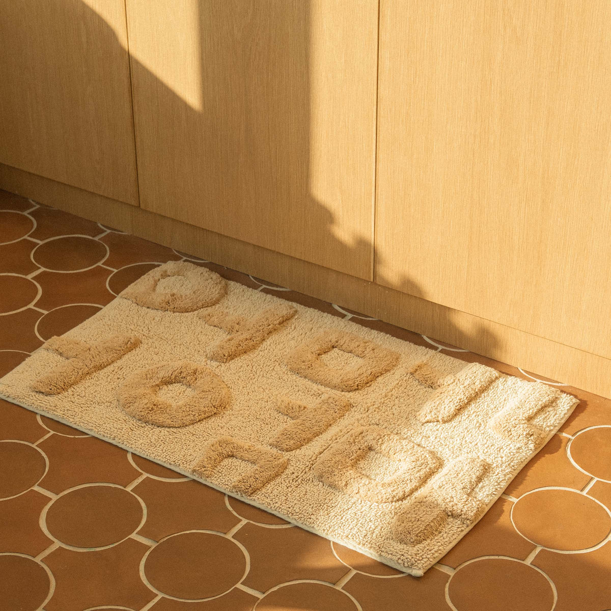 A rectangular beige bath mat with the word 'Mother' written in Korean characters, tufted in varying heights for a textured look on a tile floor in front of wooden cabinets. 