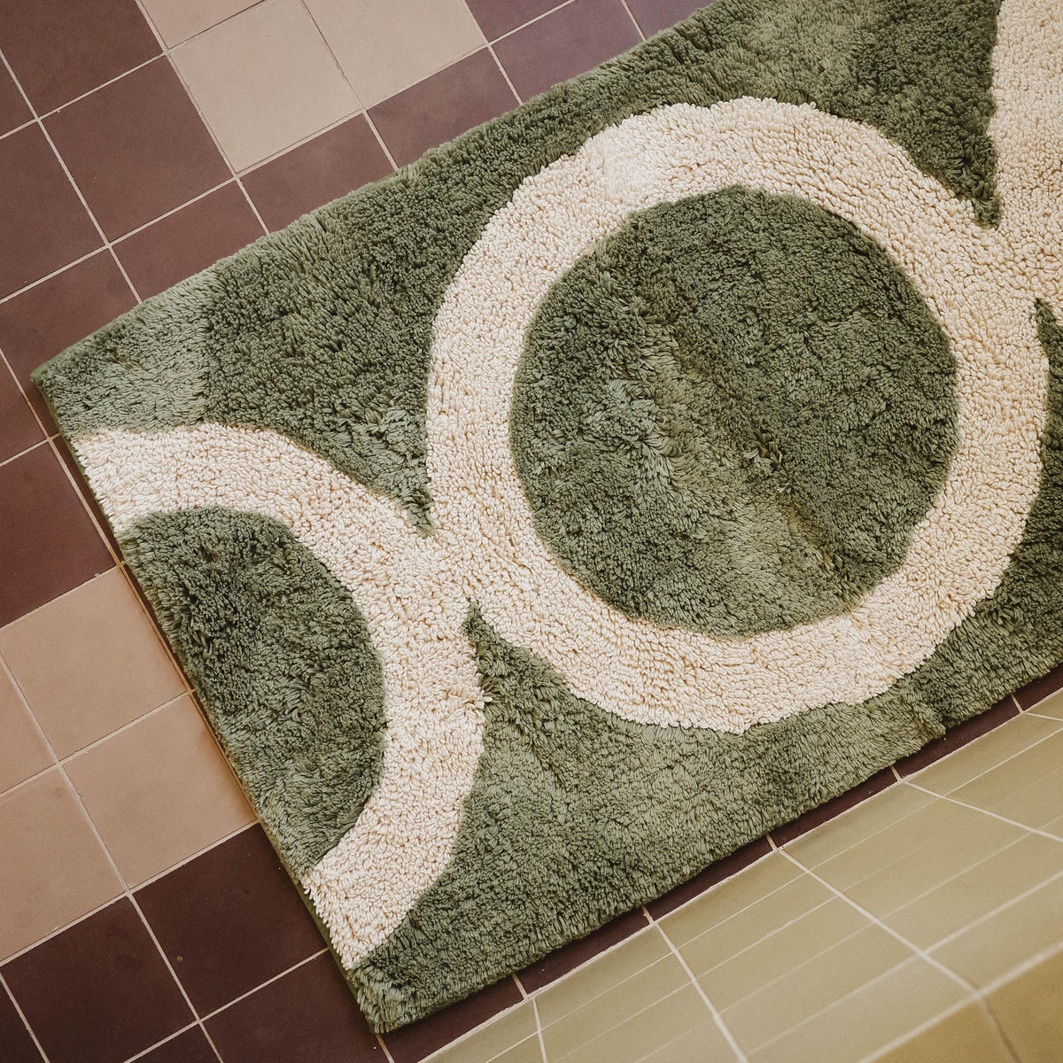 A green tufted bathmat with a beige circle pattern on a tile floor.