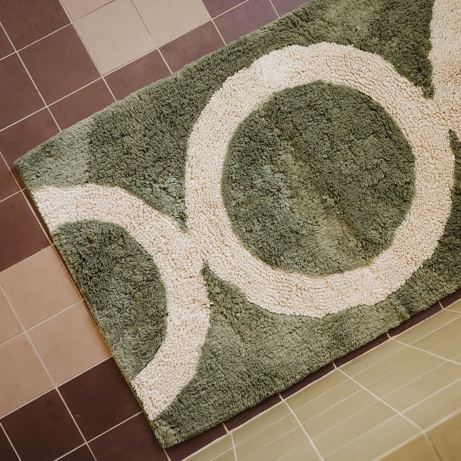 A green tufted bathmat with a beige circle pattern on a tile floor.
