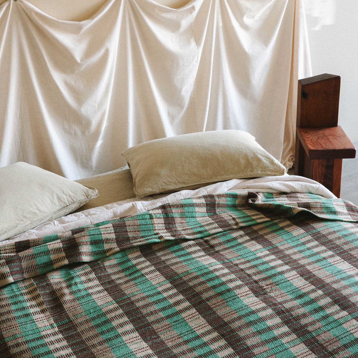 The Yara blanket on a bed with cream sheets and cream pillows against a white fabric headboard. There is a brutalist chiar in the background