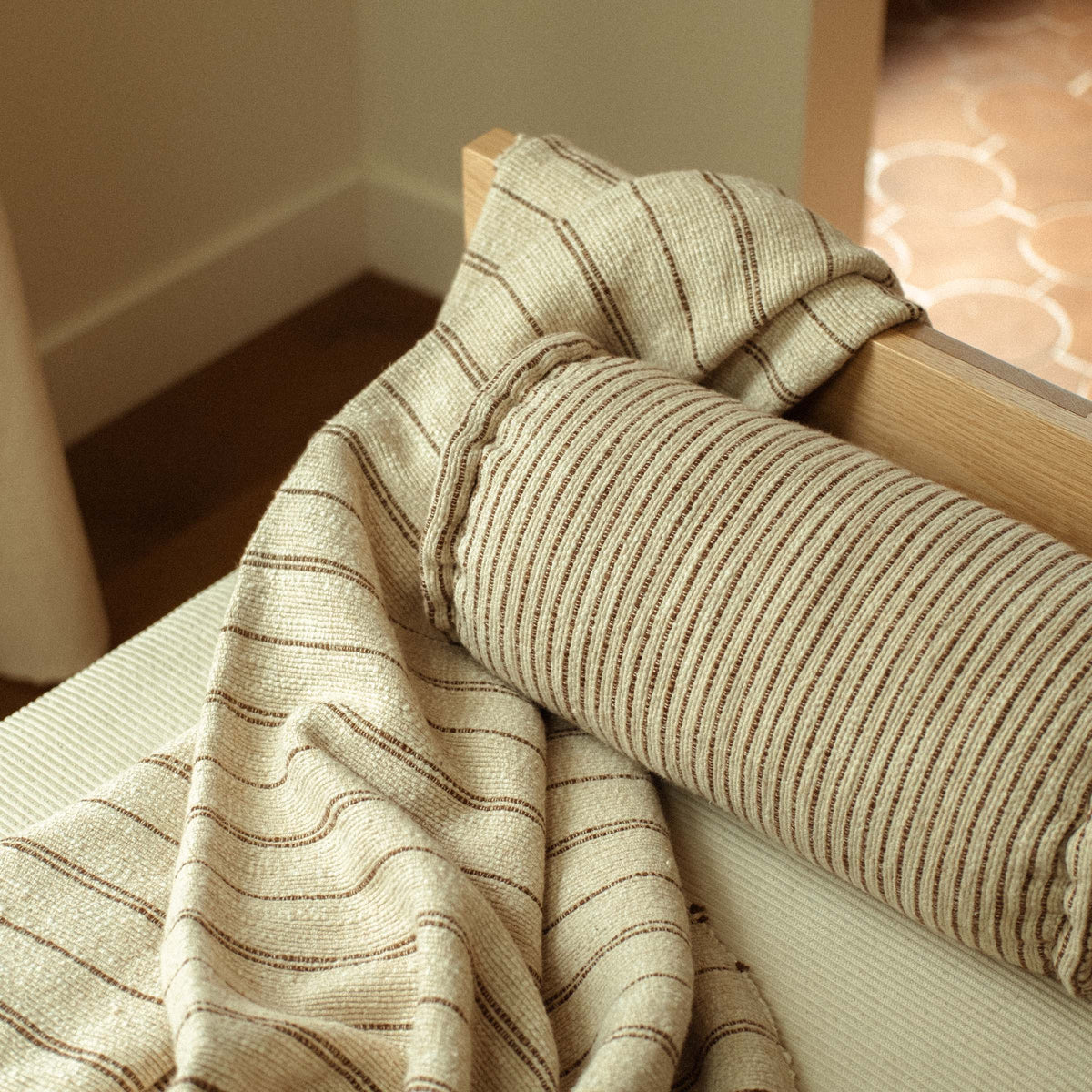A close up of the Pia blanket paired with the Pia bolster. The blanket is crumpled casually and the bolster's striper are thinner than the blanket's stripes