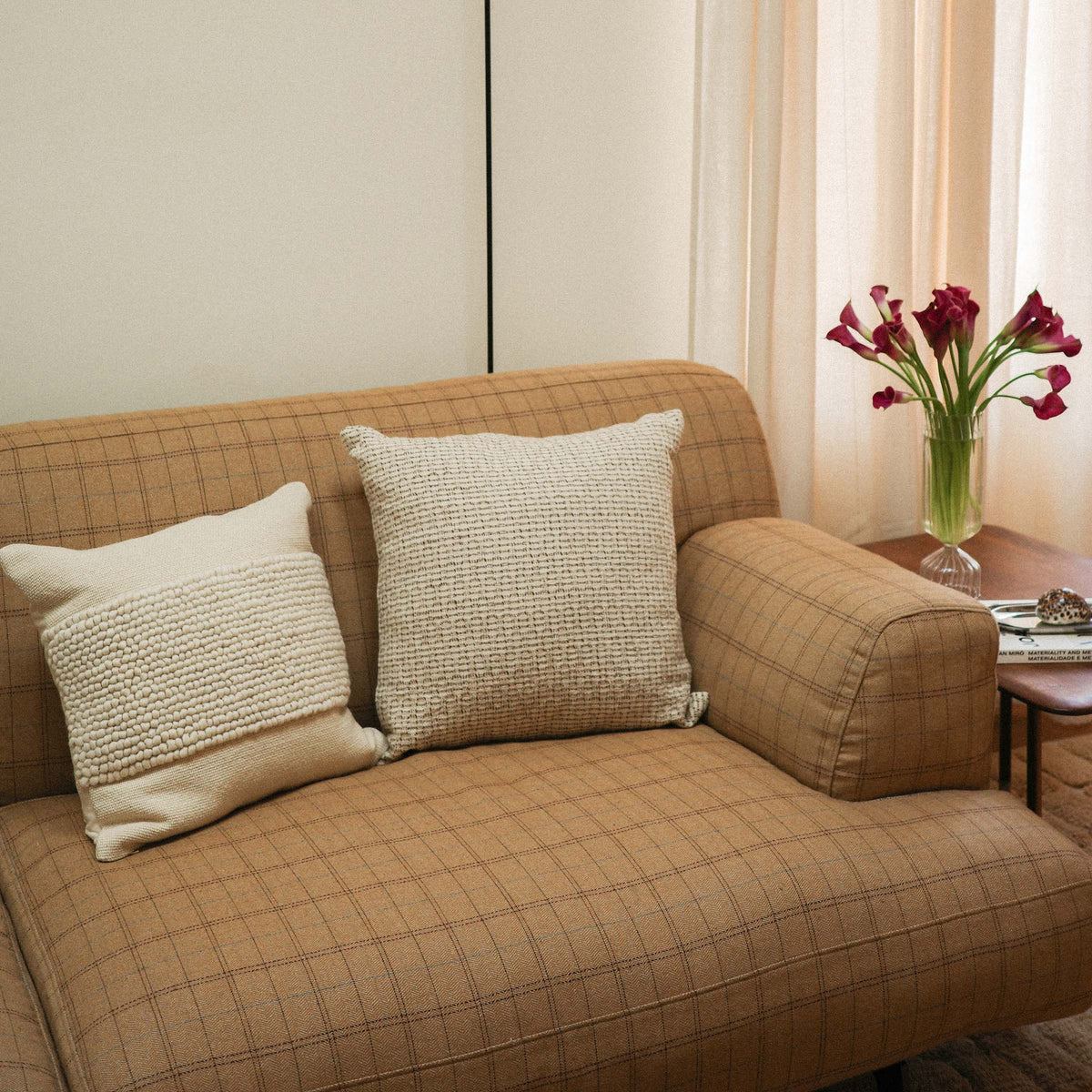A throw pillow with a beige and brown hazelnut stripe texture next to a cream colored throw pillow on a couch by a side table and flowers.