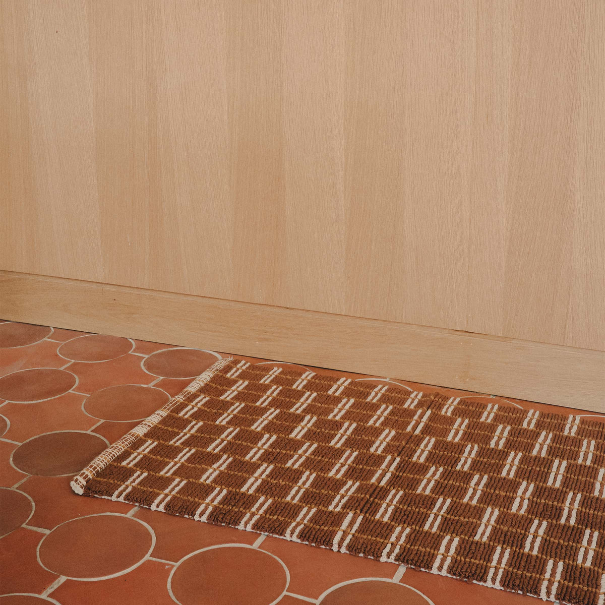 A brown bathmat with a tan and camel geometric pattern on a terracotta tile floor.