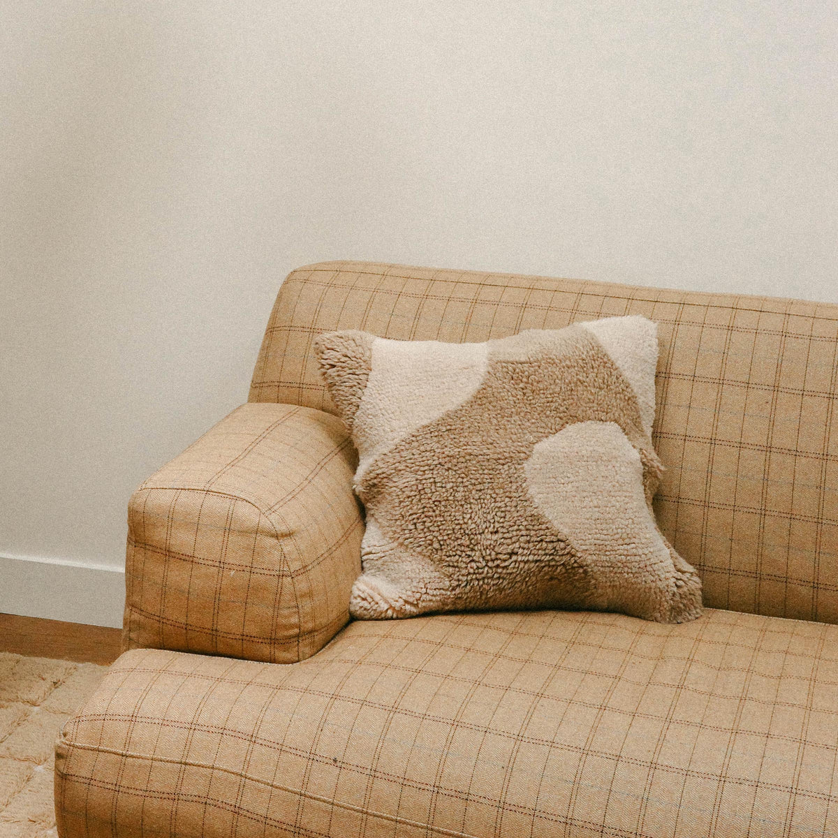 The corner of a checkered tan sofa with the Otis pillow resting. The Otis is a square throw pillow with an organic design in cream and beige.