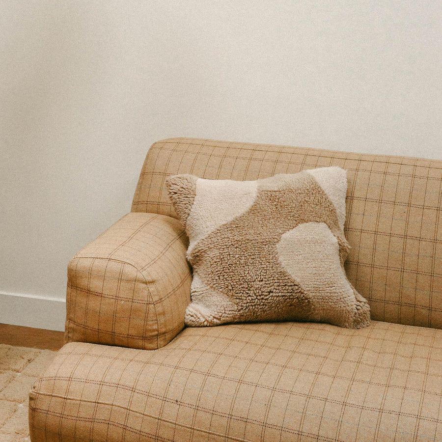 The corner of a checkered tan sofa with the Otis pillow resting. The Otis is a square throw pillow with an organic design in cream and beige.