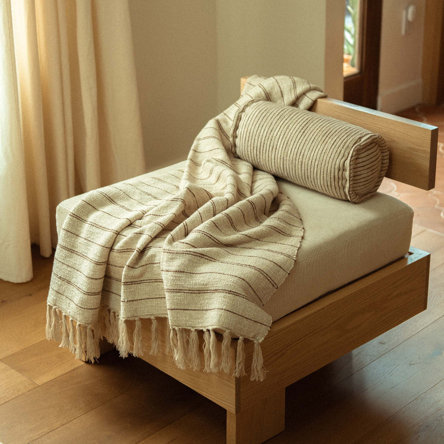 A modern accent chair featuring the Pia throw blanket which is cream and beige with stripes and tassels 