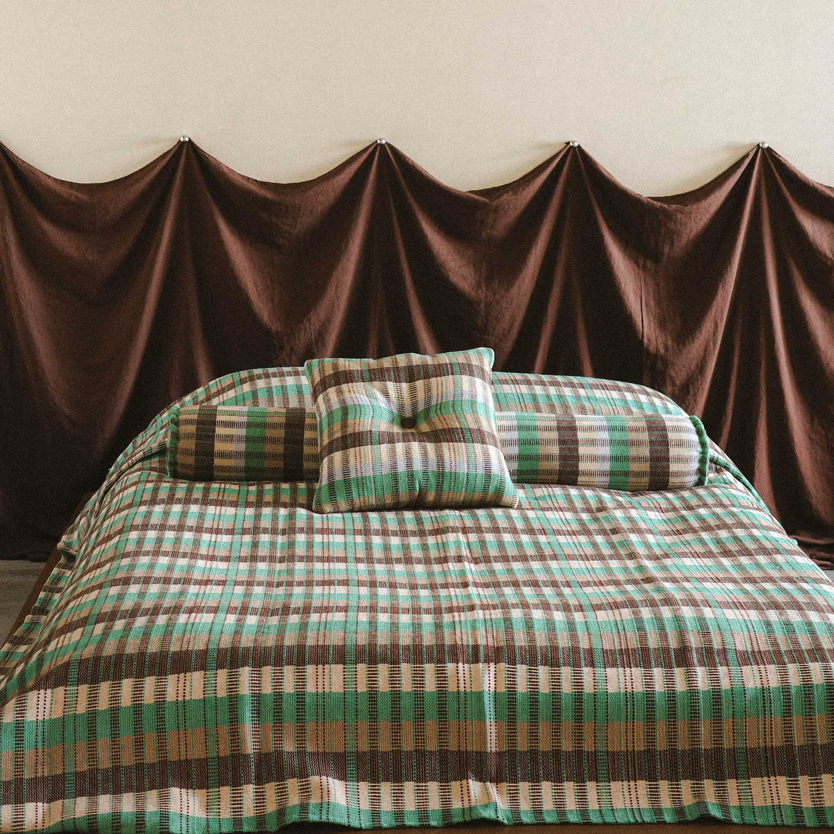 Shot of a neatly made bed with the checkered Yara bedcover on a low bed styled with the Yara bolster against a brown fabric headboard 