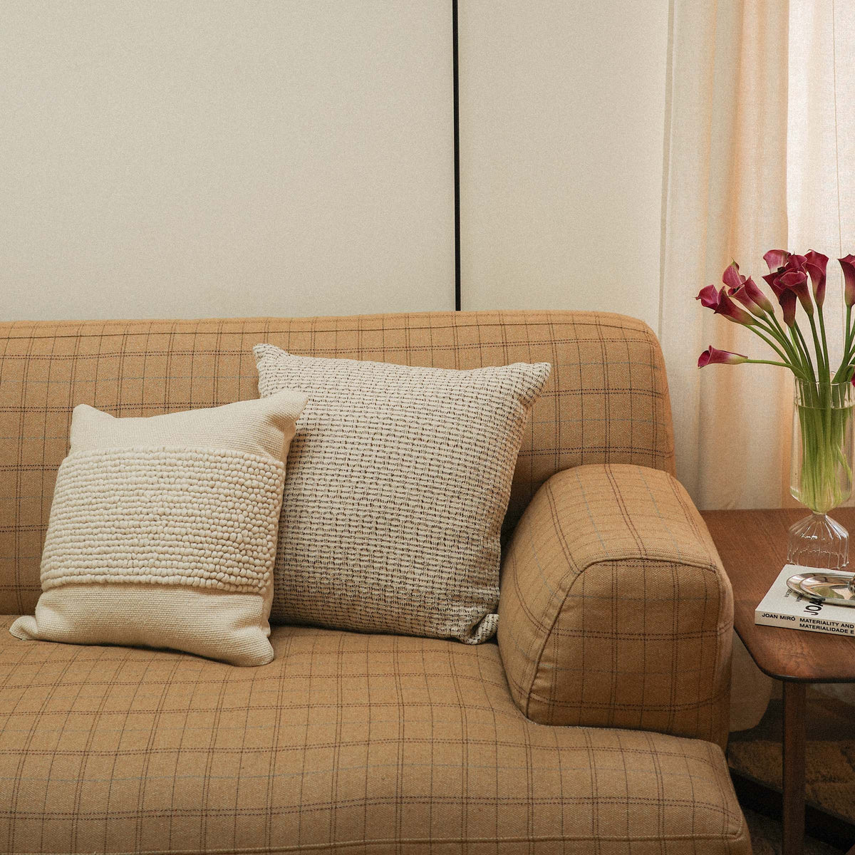 A throw pillow with a beige and brown hazelnut stripe texture next to a cream colored throw pillow on a couch by a side table and flowers.