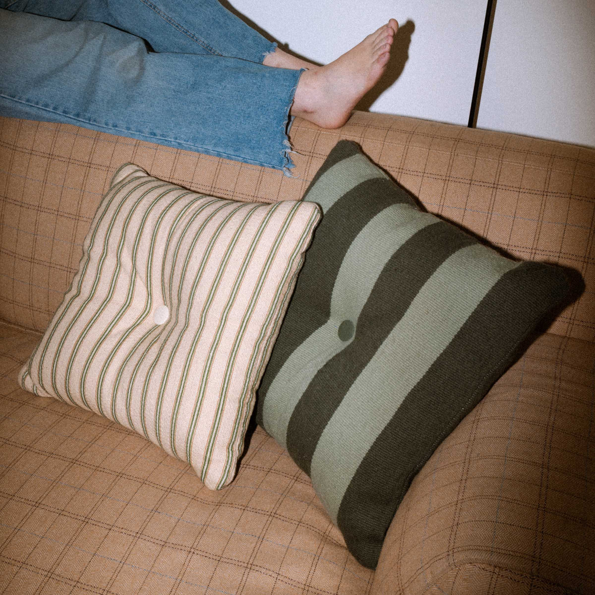 Two green striped pillows on a couch with a person wearing blue jeans in the background.