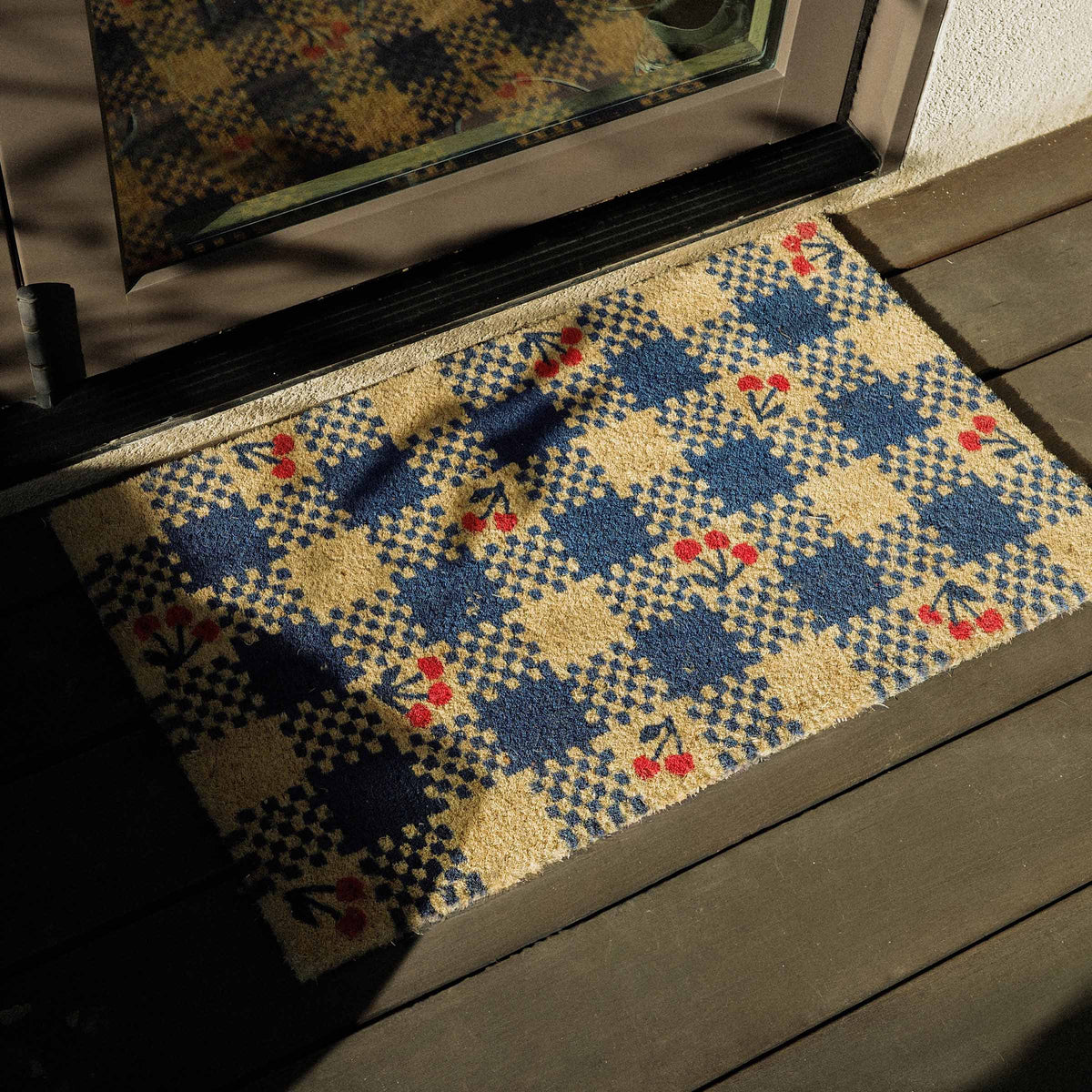 Decorative doormat with a blue geometric pattern and cherries on a wooden deck.