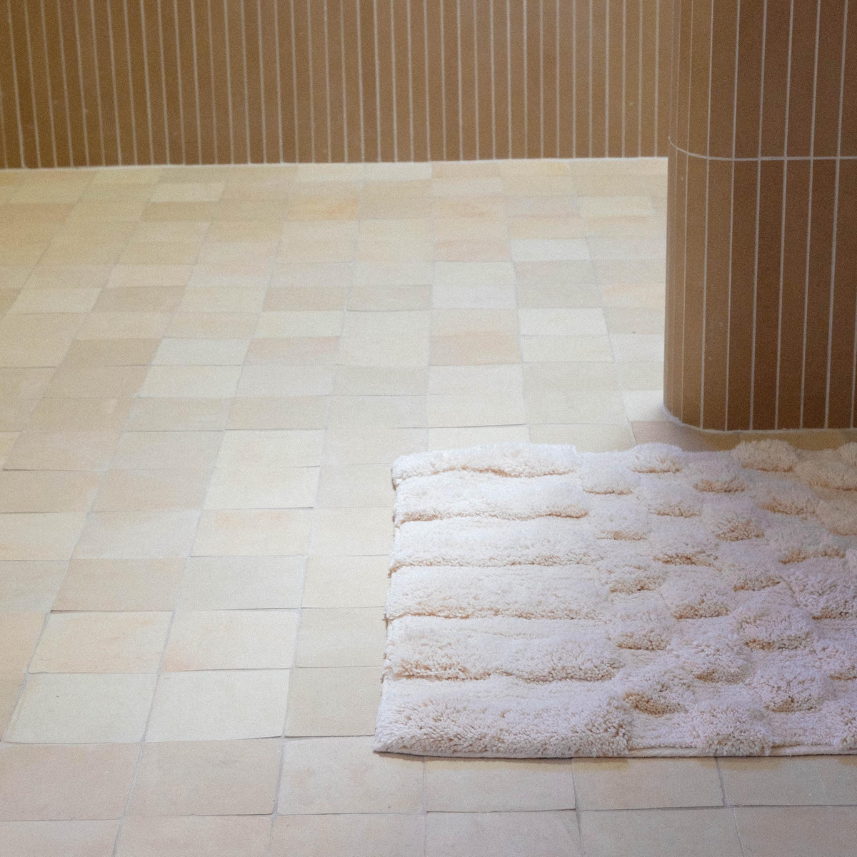 Close up of a pink bath runner with alternating shag heights in a bathroom with brown vertical tiles along the wall.