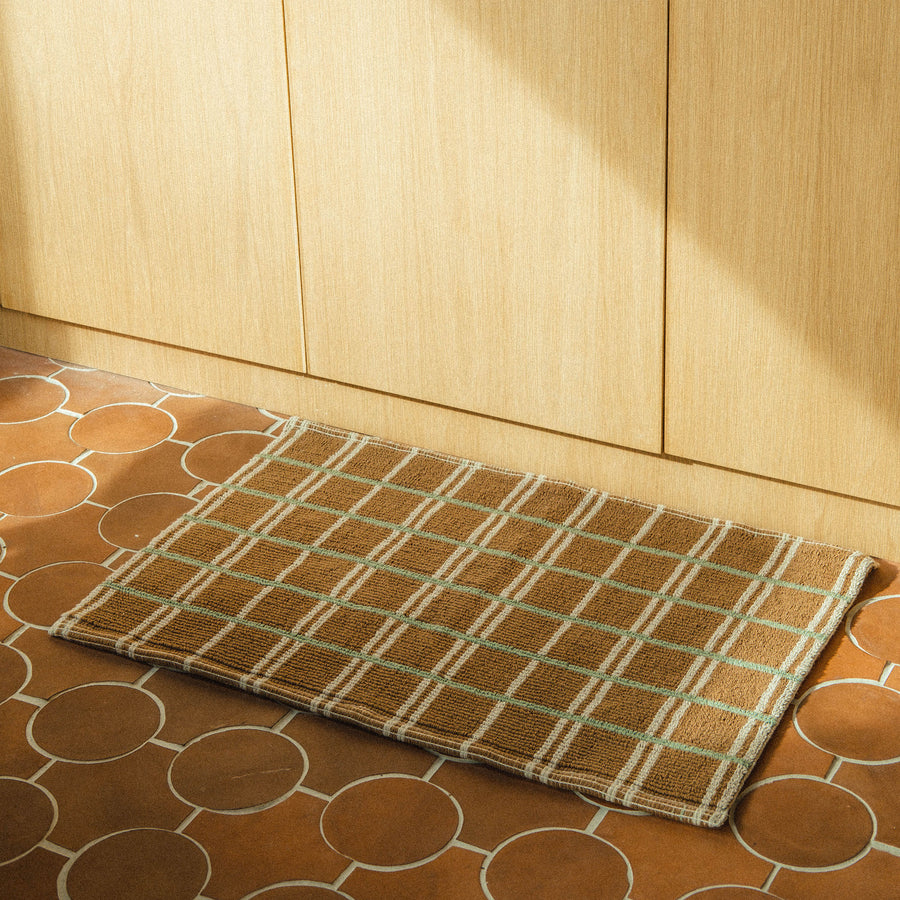 A brown mat with green and tan intersecting stripes on a terracotta tile floor of a kitchen.