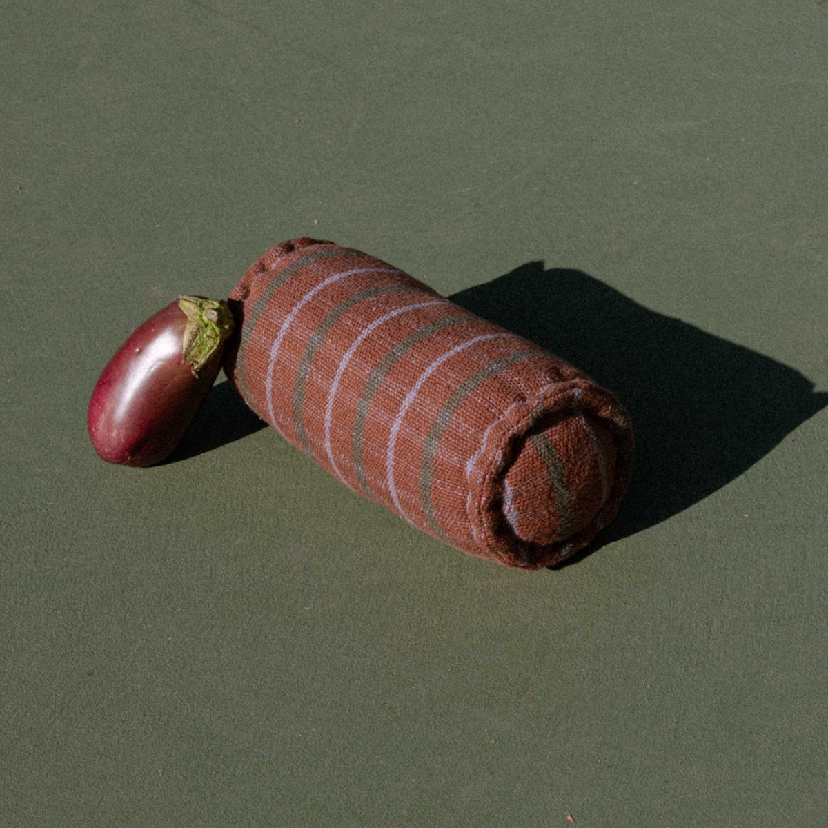 Brown cylindrical bolster featuring green and lavender check pattern on tennis court with an eggplant leaning against it.