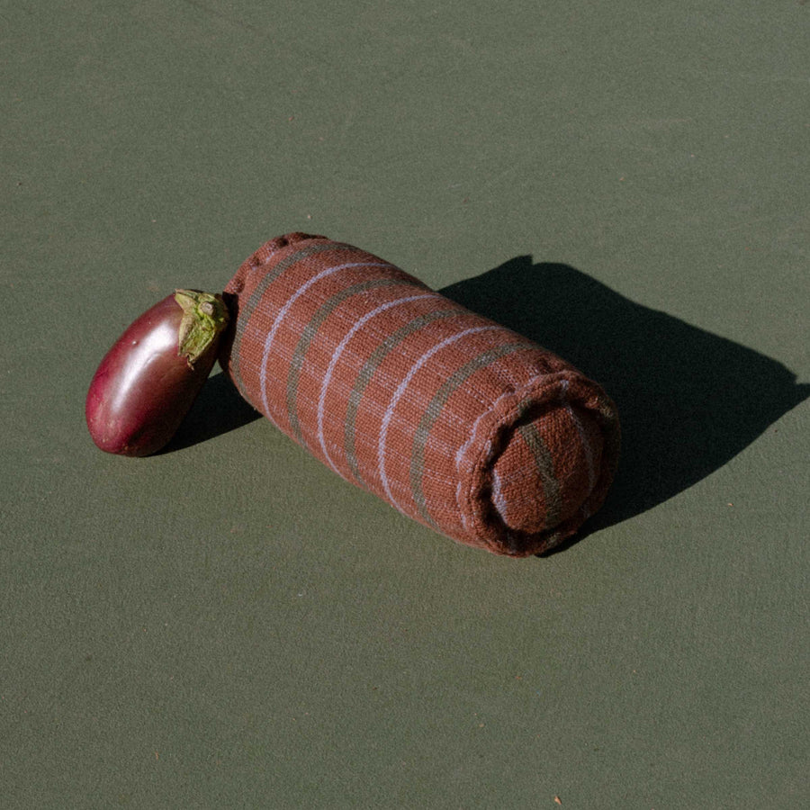 Brown cylindrical bolster featuring green and lavender check pattern on tennis court with an eggplant leaning against it.