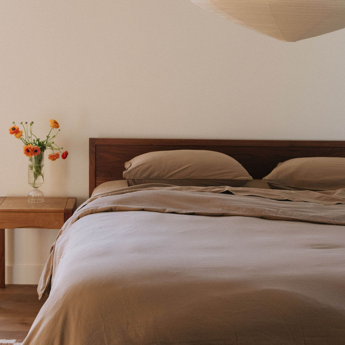 Bed dressed in beige sheets next to a nightstand with a vase of orange flowers