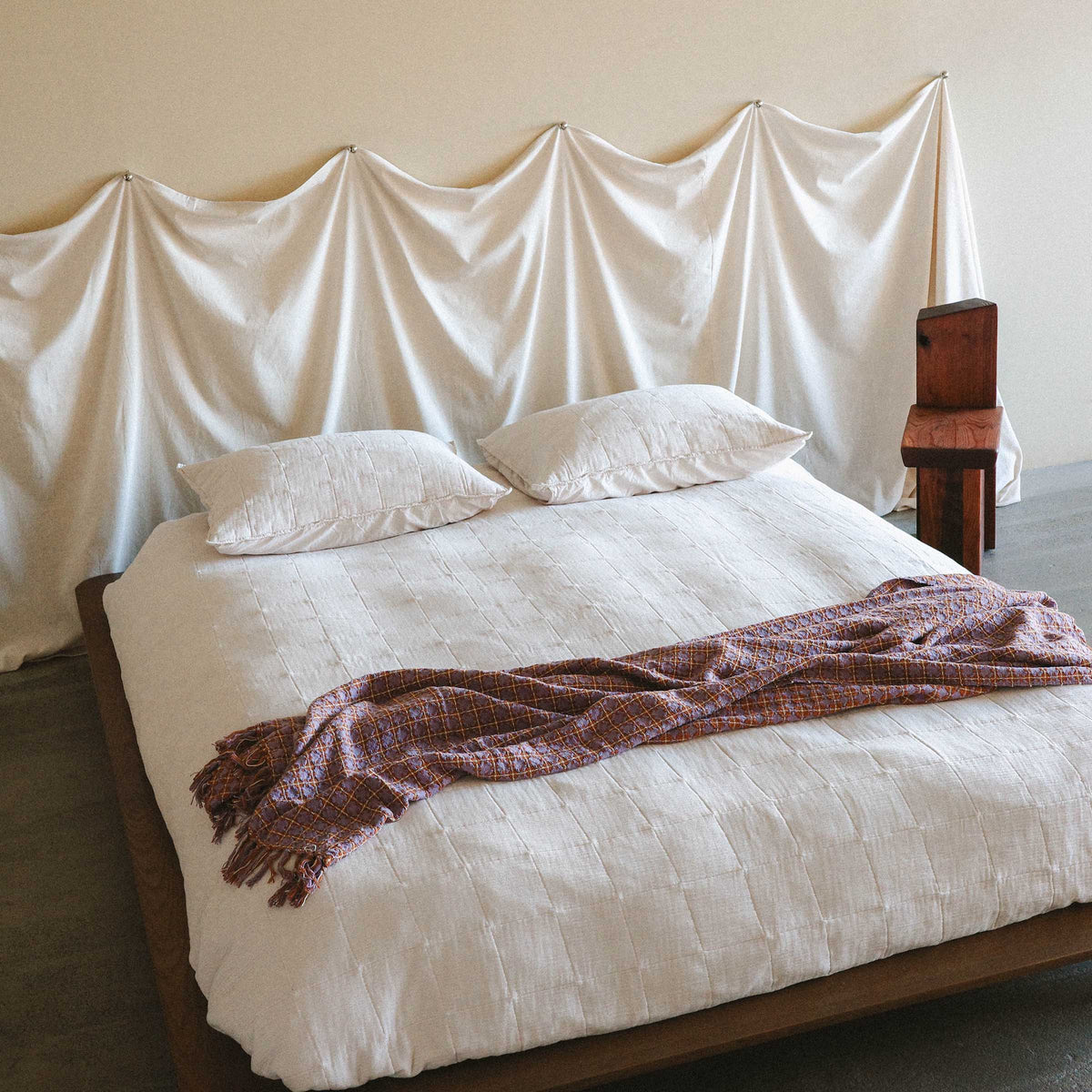 A bright bedroom setting with a white bed and pillowcases and the Rosina blanket thrown casually on the lower part of the bed. 