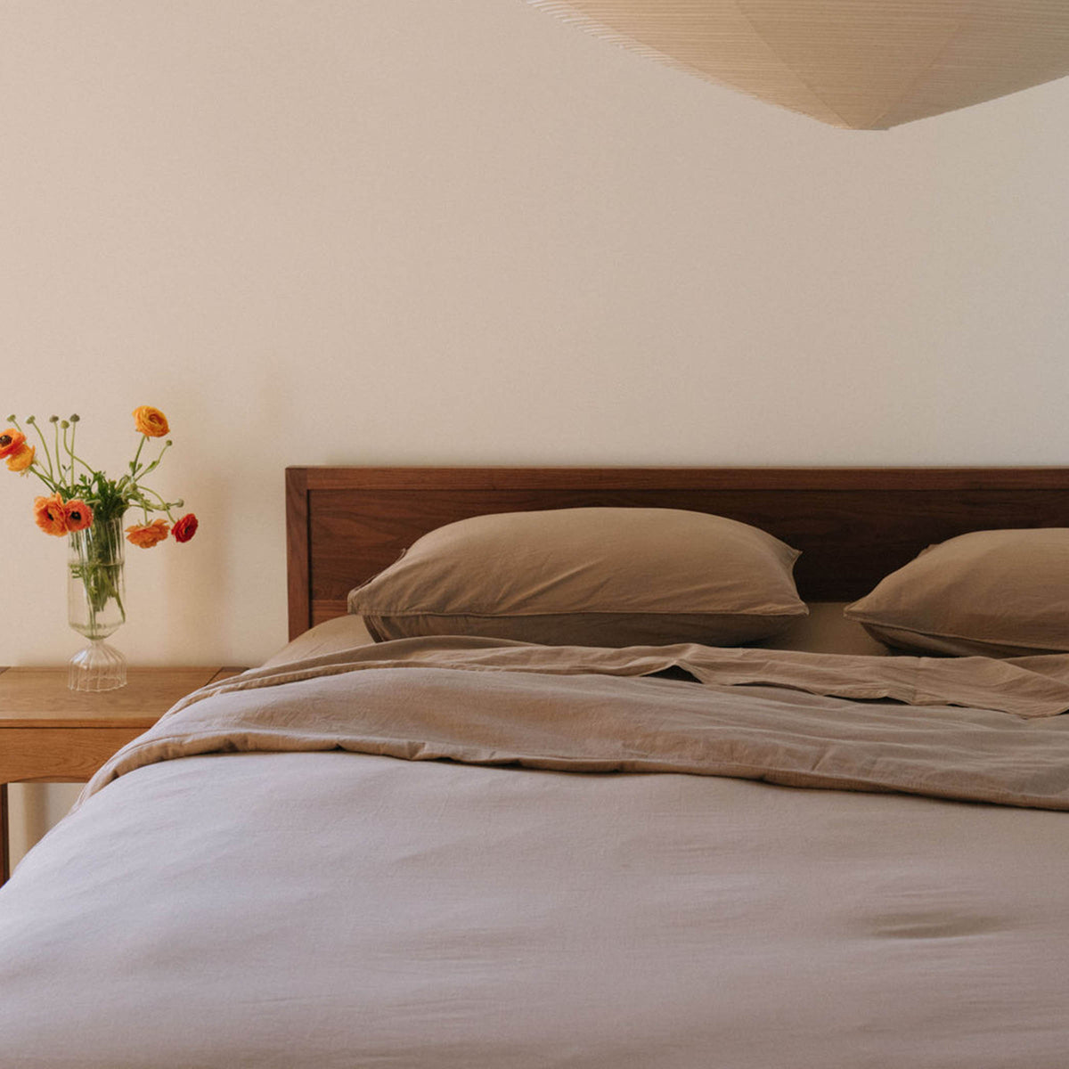 A bed dressed in beige sheets with two pillows against a wooden headboard and next to a nightstand with a vase of flowers