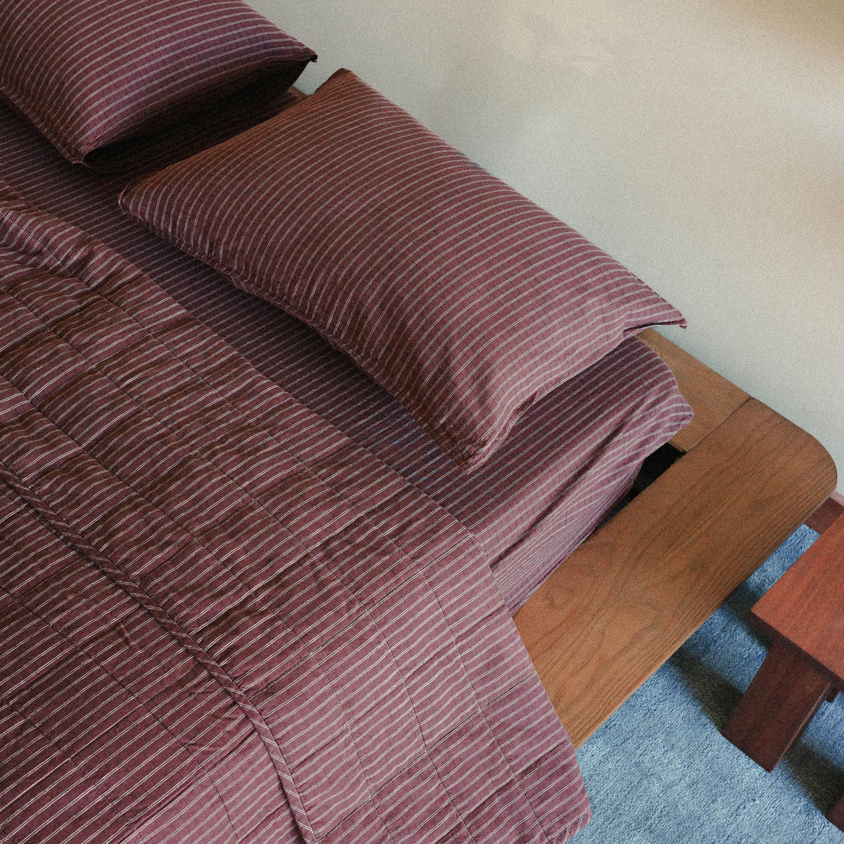 A close up of the top of the bed with a brown / maroon striped quilt / bed cover, sheets and pillows on a wooden bed frame in a bedroom setting