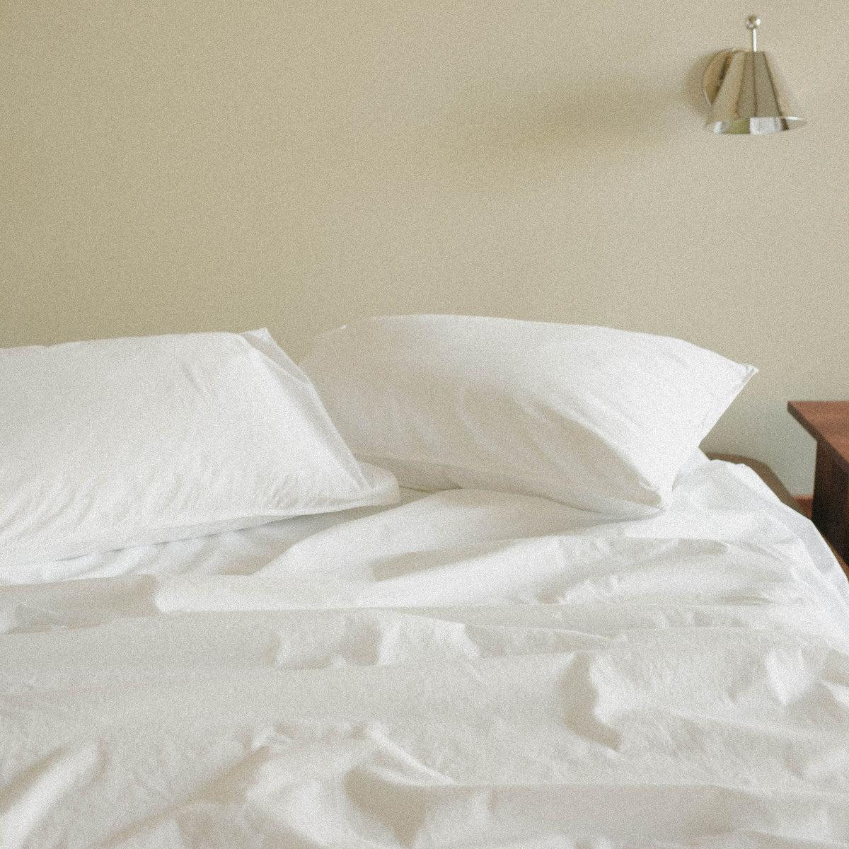 White bedding on a bed with a neutral wall and lamp in the background
