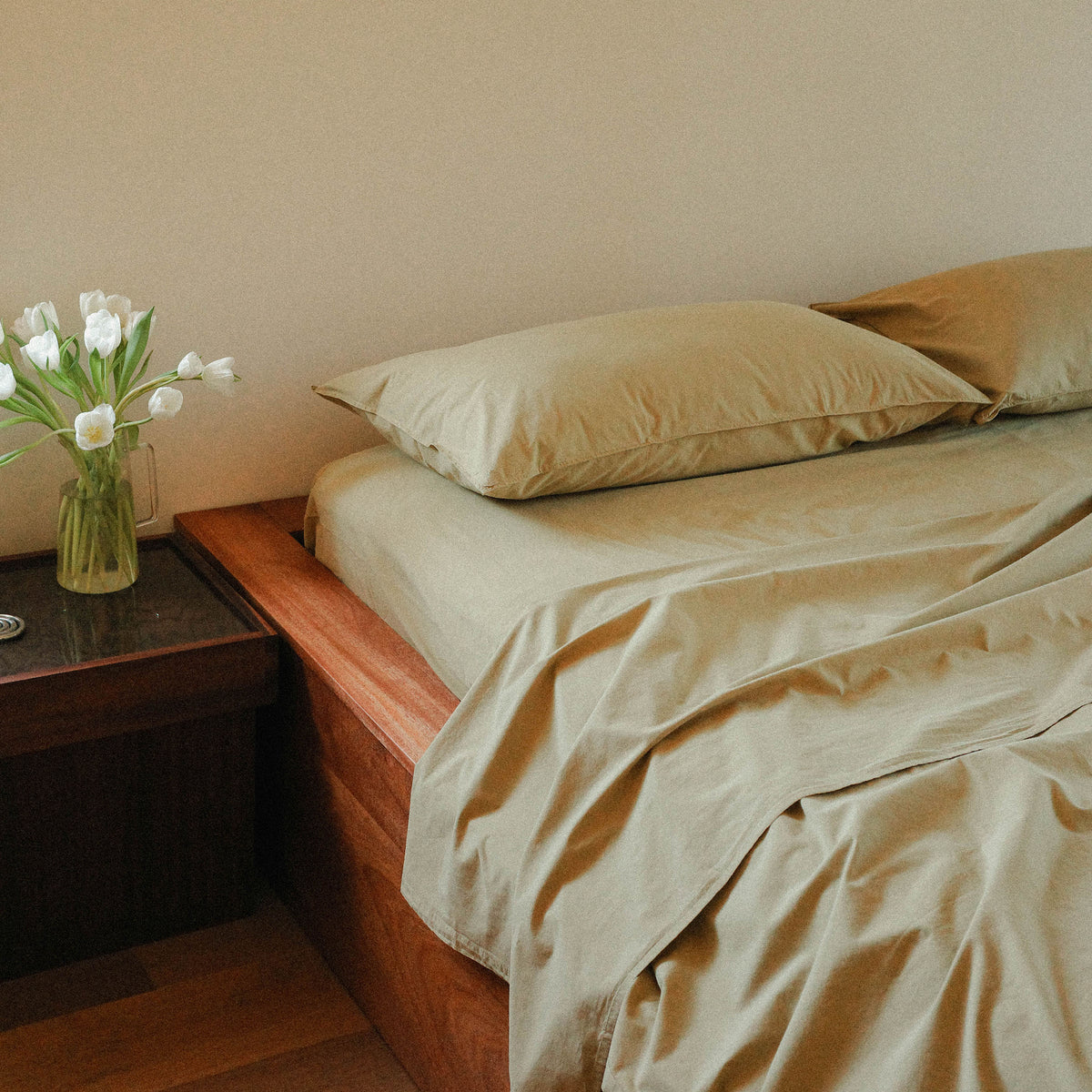 Bed with green bedding and pillows, next to a wooden nightstand with a vase of white flowers.