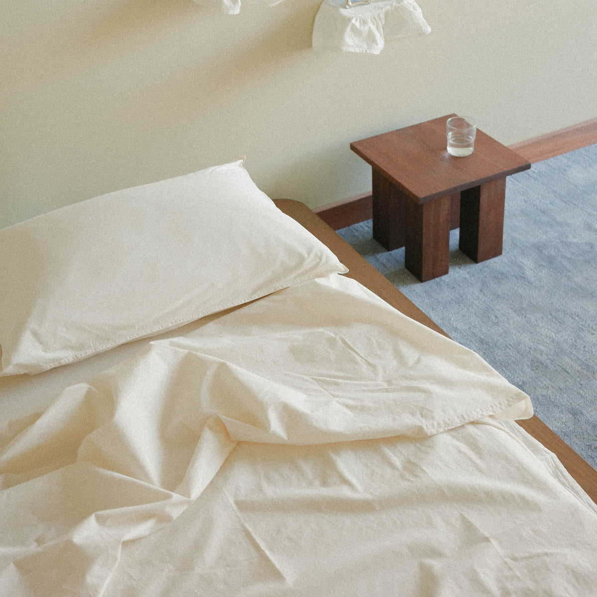 Cream sheets on a bed with a wooden nightstand and glass in a room with blue carpet.
