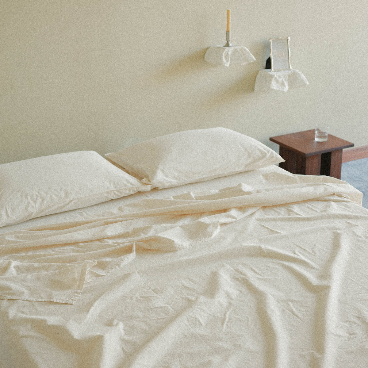 Cream sheets and pillows on a bed with a nightstand and  two floating shelves holding a taper candle and picture frame in the background.