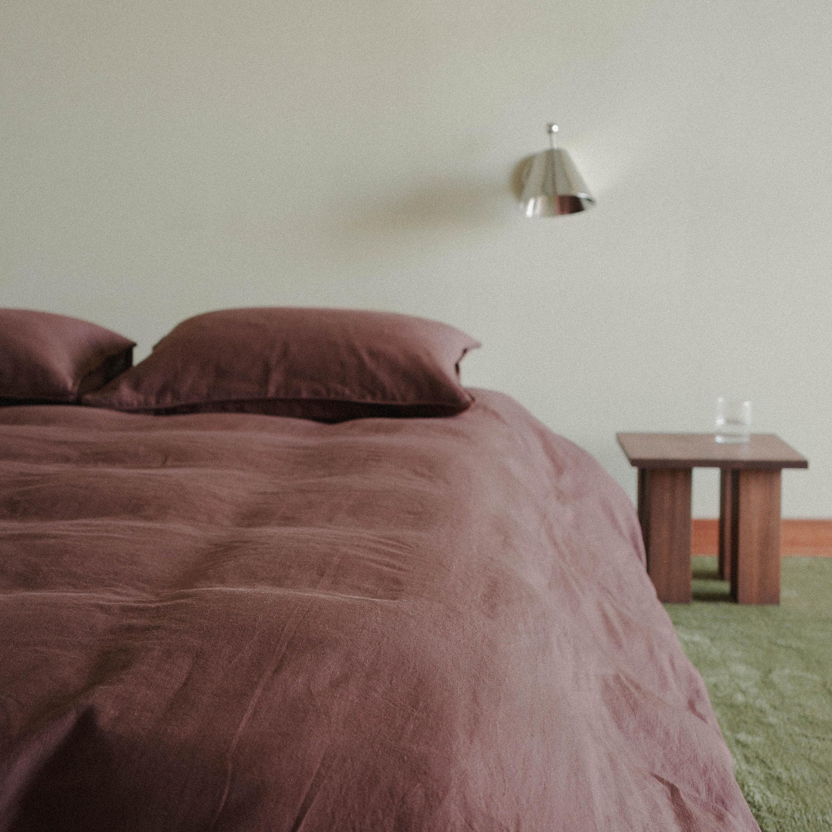A closer photo of the bed with a brown / purple duvet cover, the room is tastefully sparse with a green carpet and a wood nightstand 