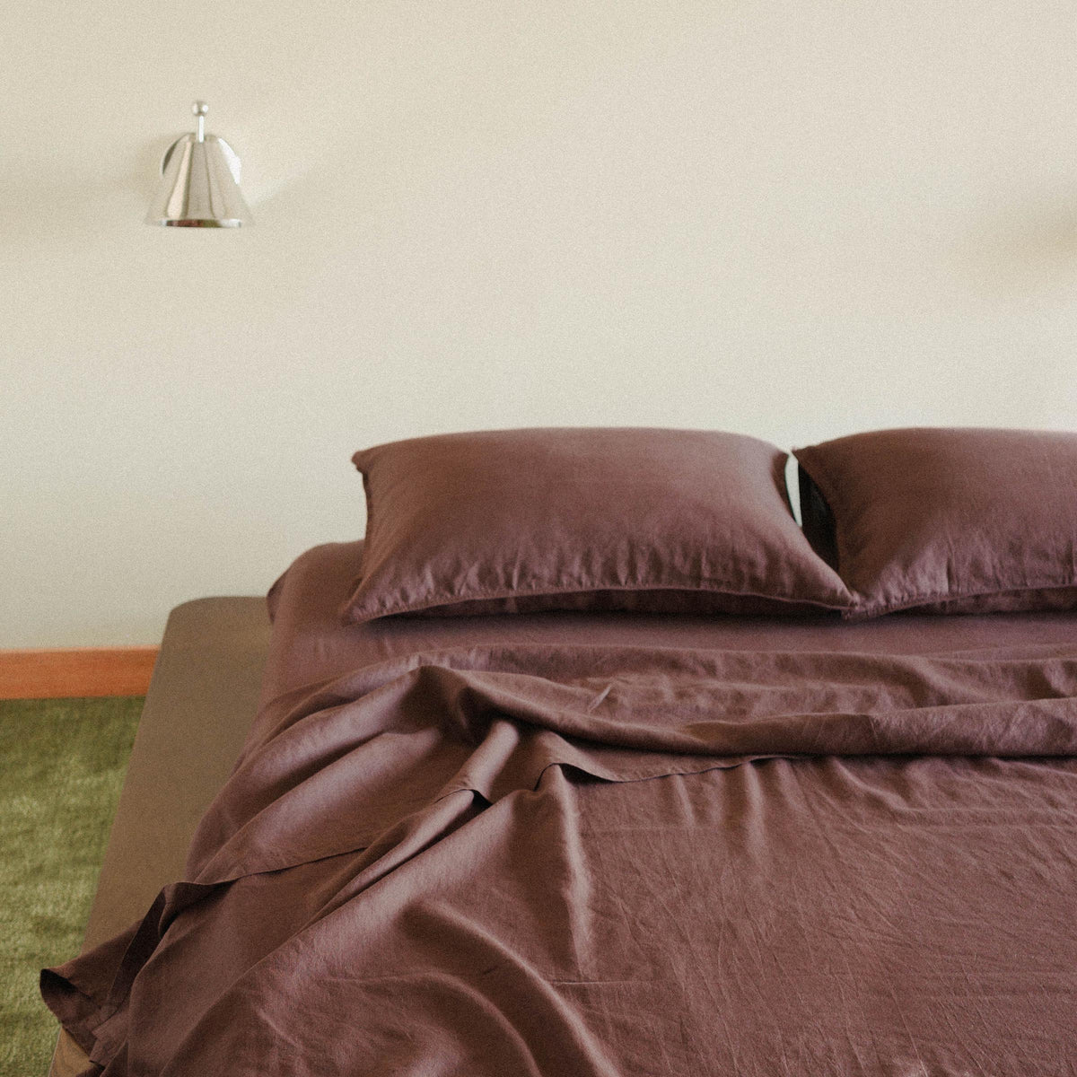 Half a bed dressed in brown / purple sheets with two pillows in a bedroom setting with a green carpet 