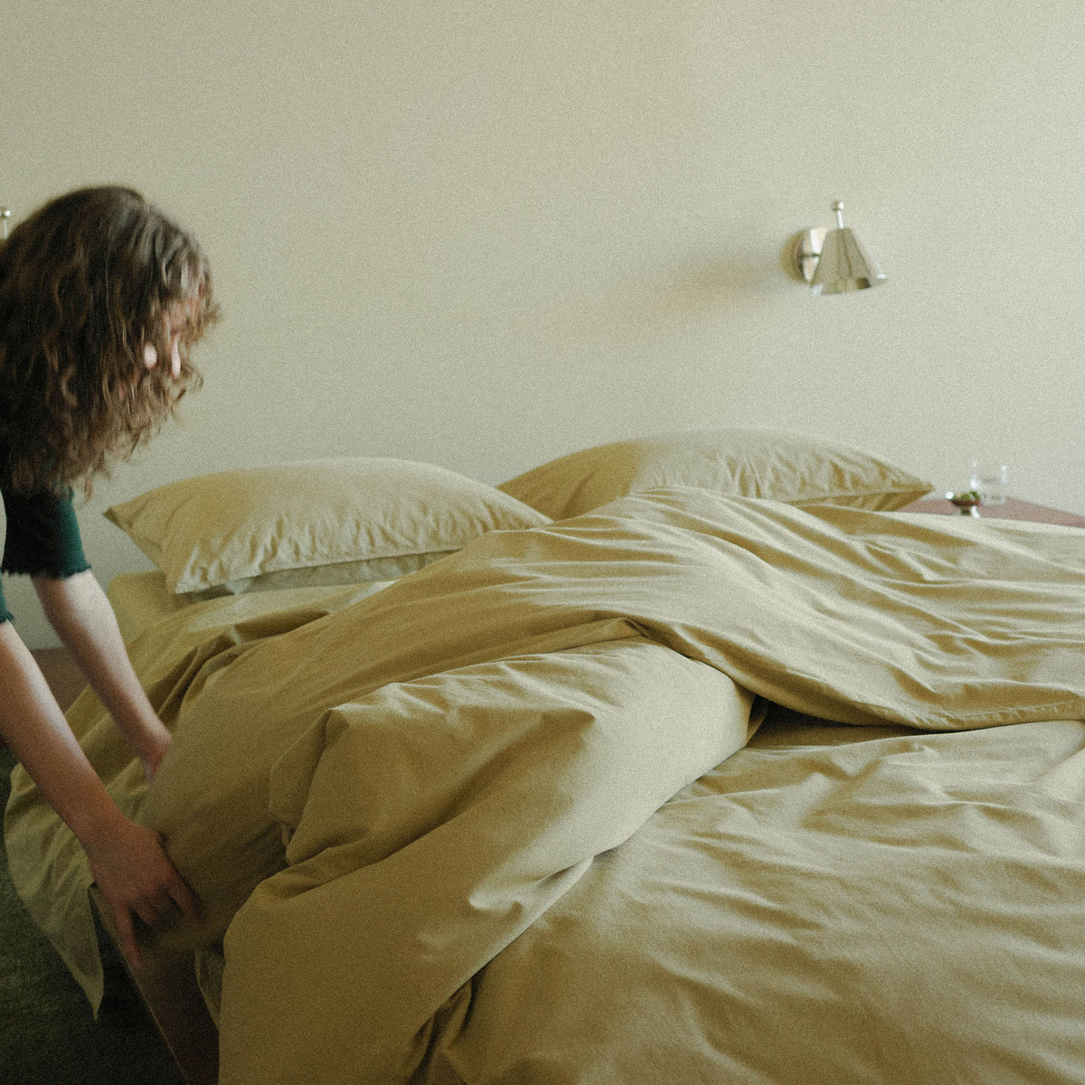 Person making a bed with green bedding in a room with a light-colored wall and a silver wall sconce.