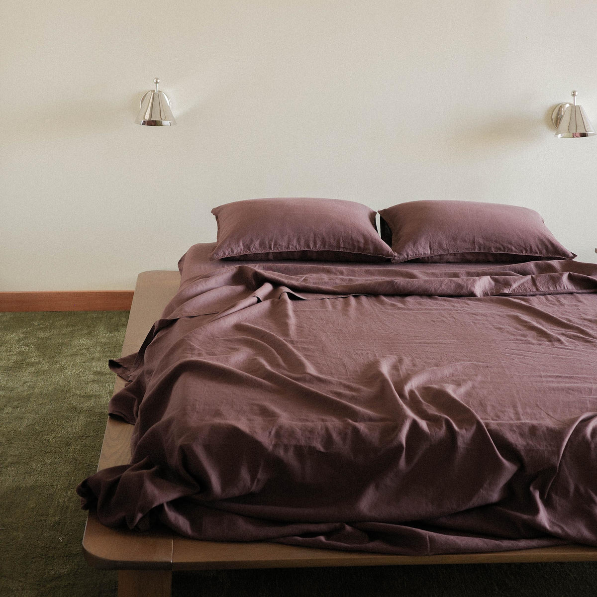 A bed dressed in brown / purple sheets, two pillows. The bed is a wooden platform bed in a bedroom setting against a green carpet. 