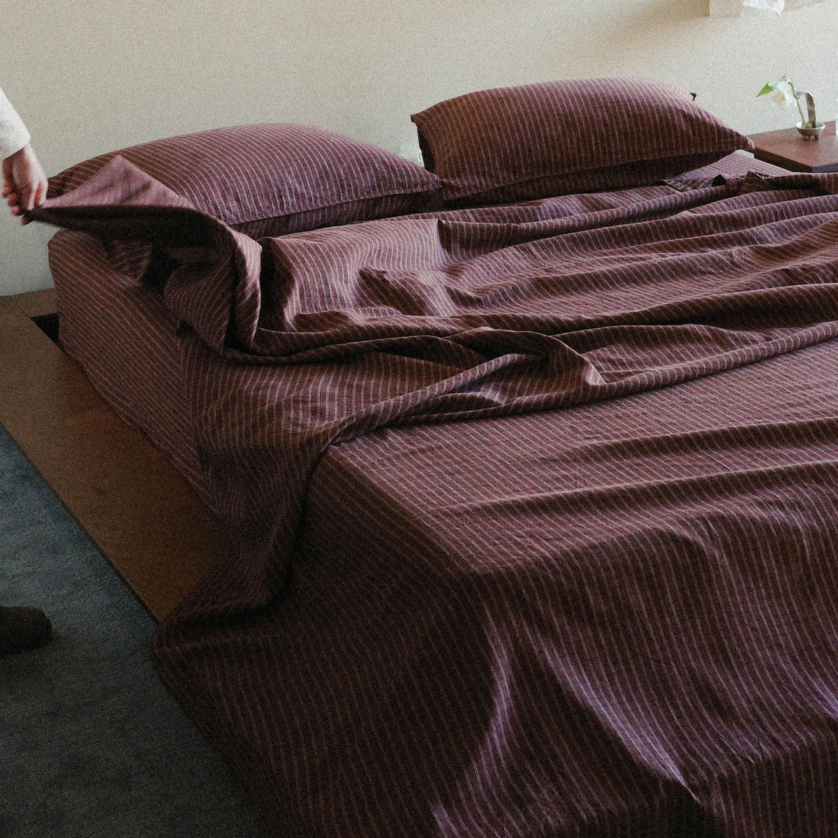 Brown striped bedspread on a bed with pillows in a bedroom setting with a hand pulling a corner of the sheet