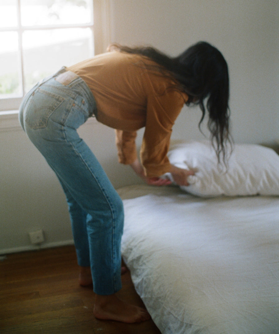 A woman bends over a bed and tucks white bedding behind a white pillow, wearing a camel long-sleeve top and light blue jeans in a softly lit bedroom.