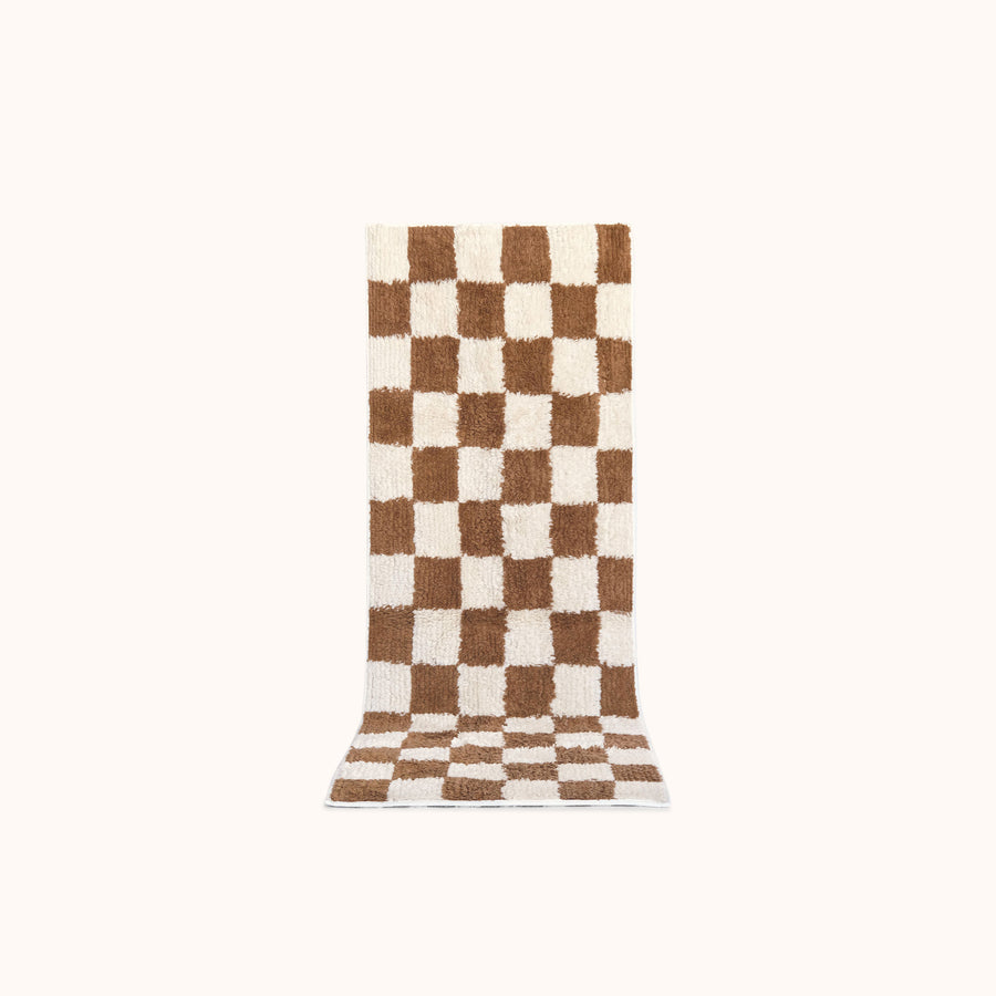A long rectangular plush bath mat with a brown and off-white checkerboard pattern, shown flat against a solid off-white background.