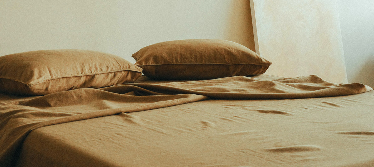 A close-up of a bed made with matching green-brown bedding, including two pillows and a rumpled sheet, set against a neutral beige wall.