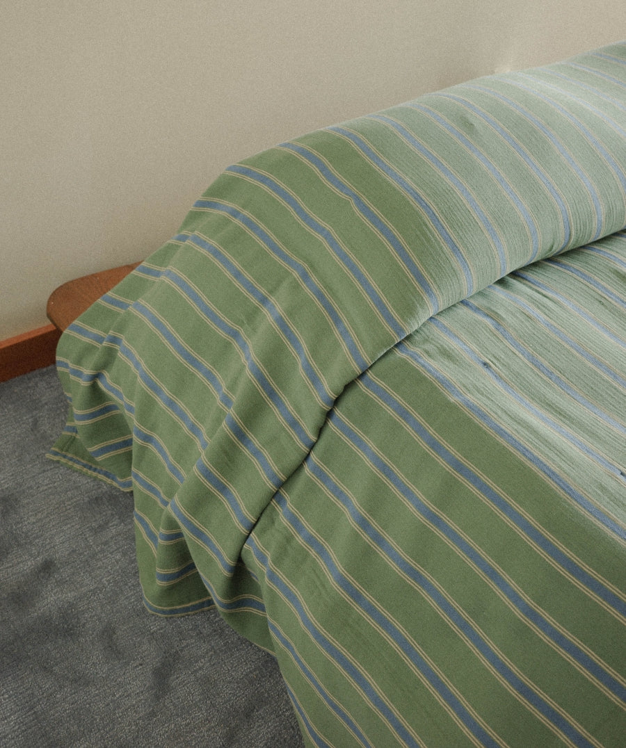 Green striped bedding, including a pillowcase and duvet cover, draped over the corner of a bed in a bedroom setting.